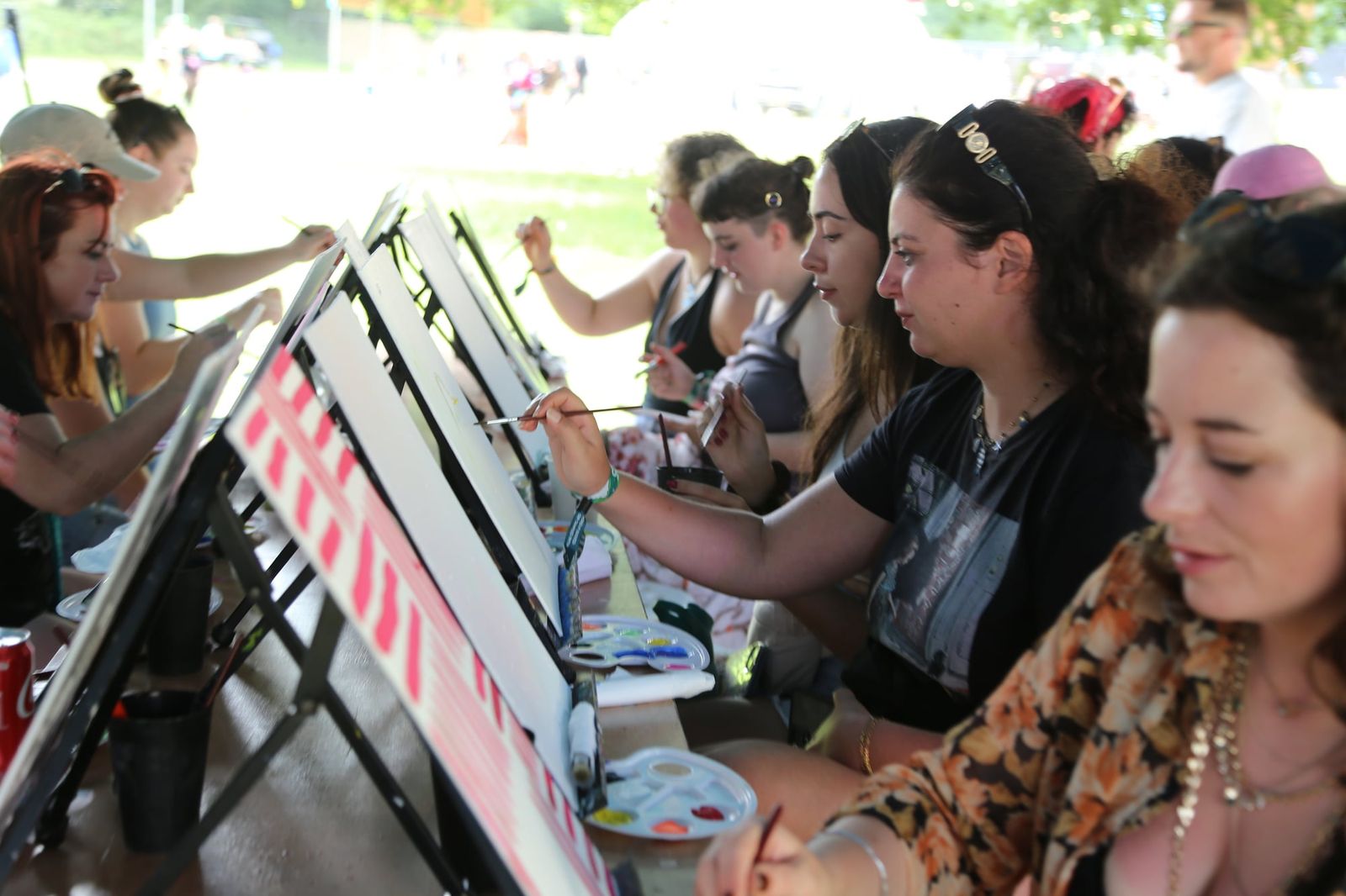 Row of guests painting at easels under a canopy at an outdoor paint event
