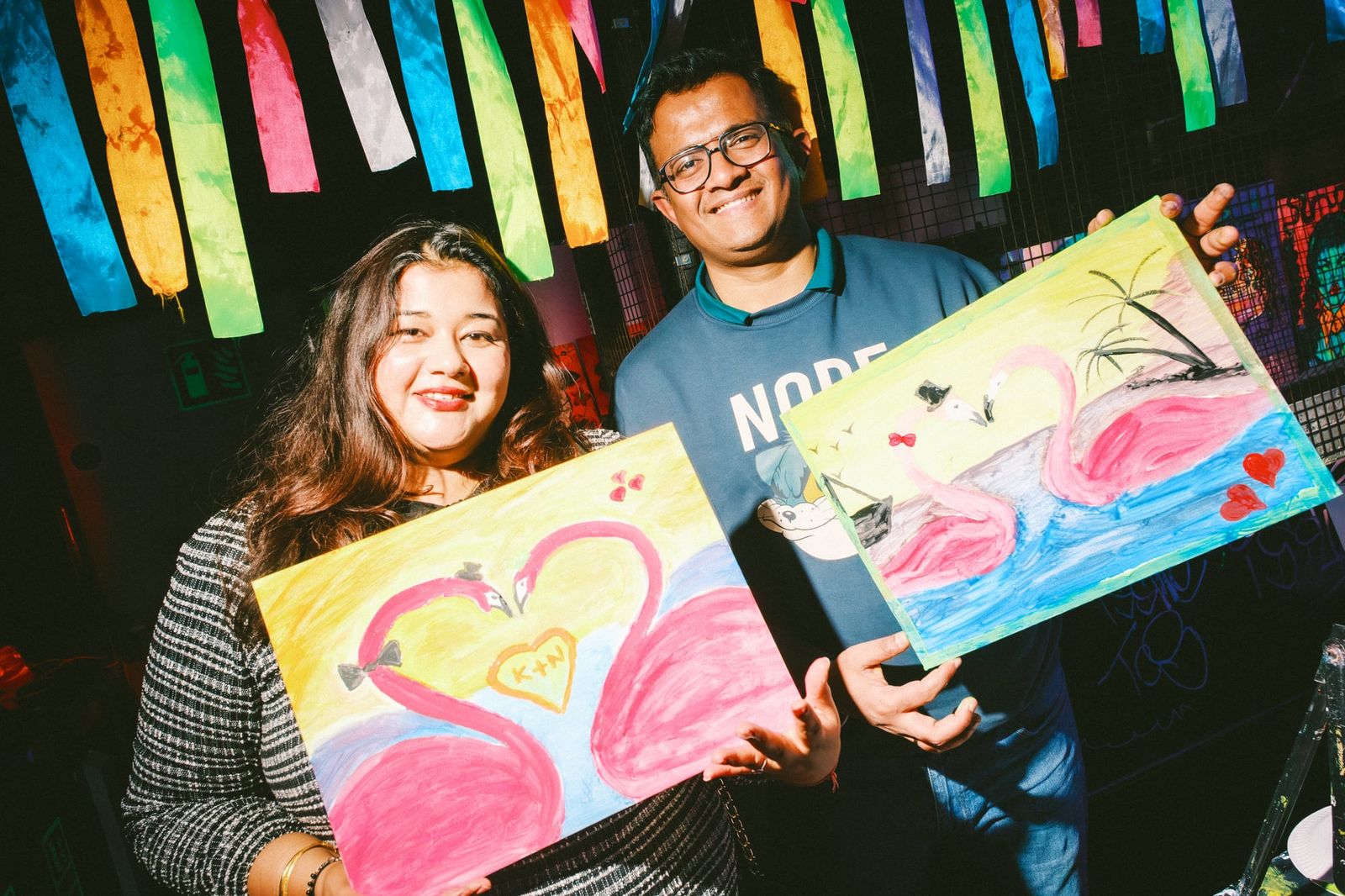 Two guests proudly holding up their matching flamingo paintings at a neon workshop