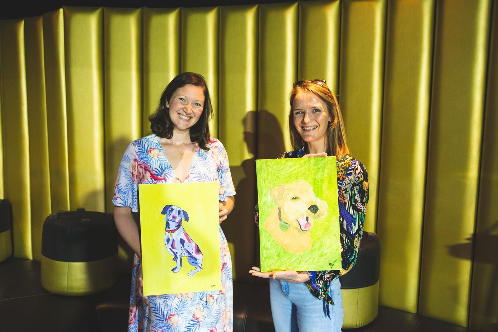 Two guests smiling while holding finished dog portrait paintings at a paint and sip event